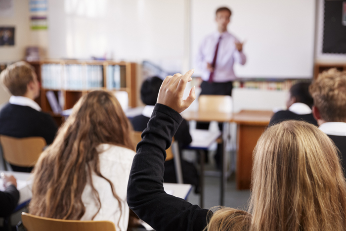 View from behind of classroom with a female pupil holding hand up to ask a question